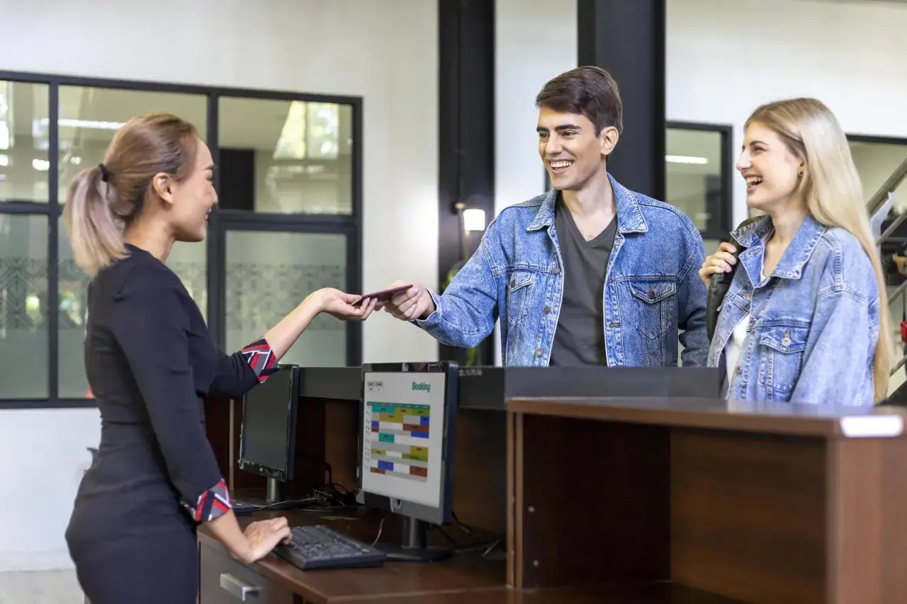 Receptionist consegna un passaporto a una coppia sorridente. Un corso di inglese per receptionist potrebbe aiutarli a comunicare meglio.