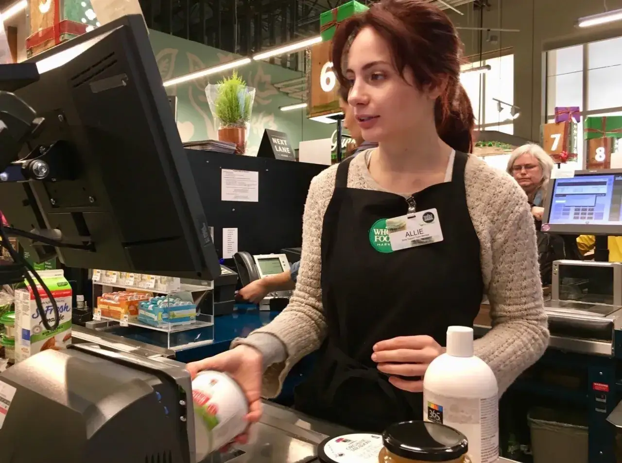 Cassiera al supermercato, Allie, con il suo badge, mentre scansiona un prodotto. Il suo stipendio da cassiera supermercato &egrave; meritato.