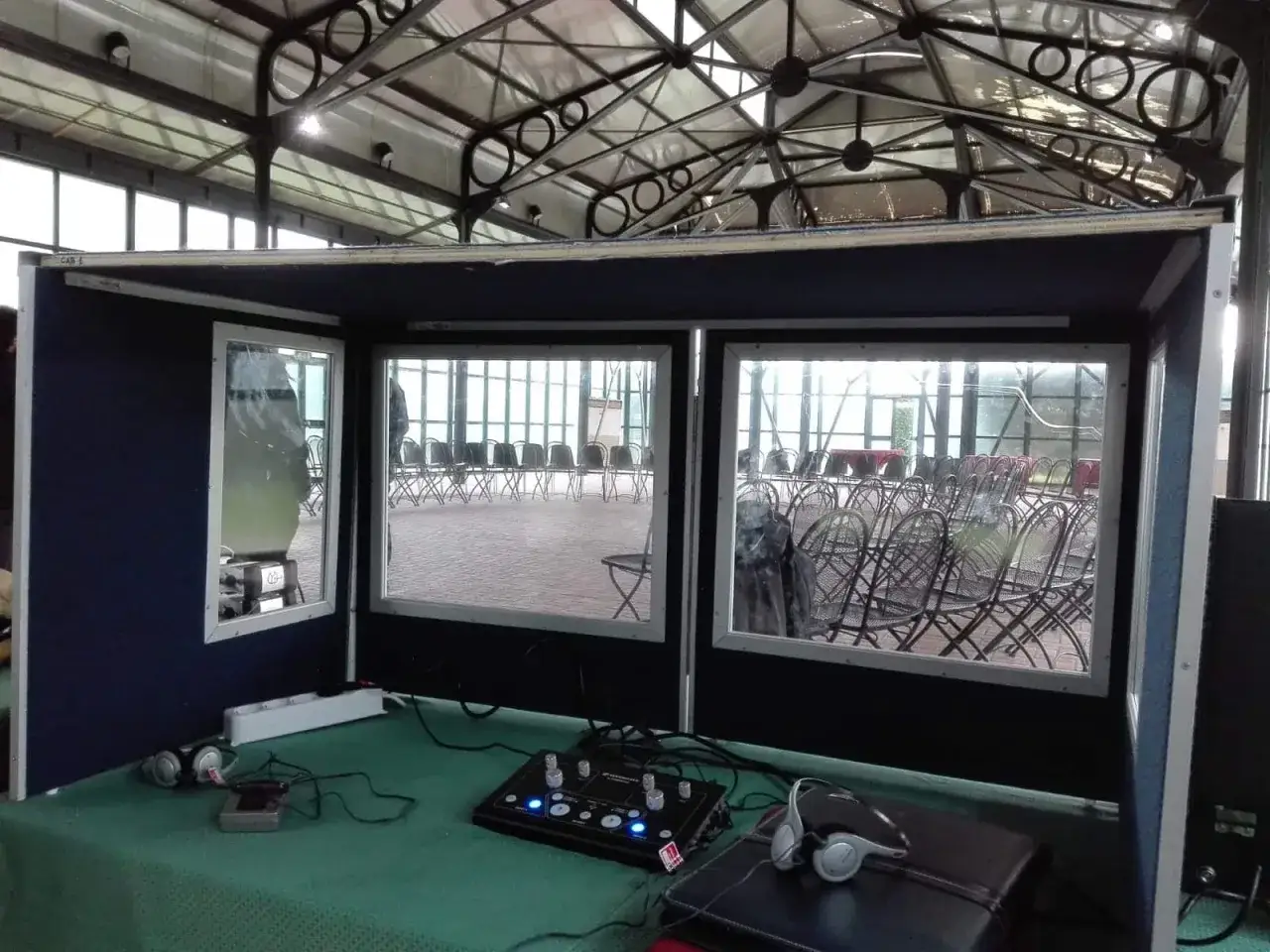 Cabina per interpretariato di conferenza con cuffie e console, vista su una sala con sedie disposte.