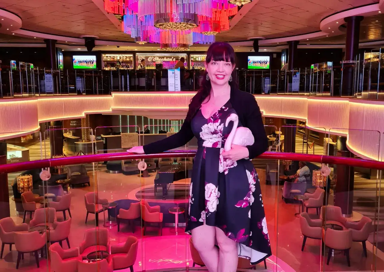 Donna sorridente con fenicottero rosa, felice di lavorare sulle navi da crociera. L'ambiente &egrave; un lounge elegante con luci colorate e arredi moderni.