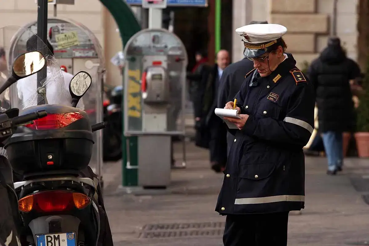 Vigile urbano in divisa scrive su un taccuino, forse annotando dettagli per il suo stipendio. Sullo sfondo, cabine telefoniche e un motorino.