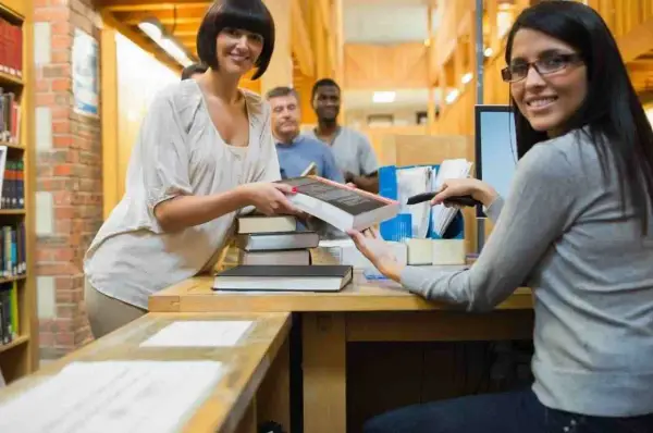 Due donne sorridono mentre lavorano in biblioteca, una passa un libro, l'altra usa uno scanner.